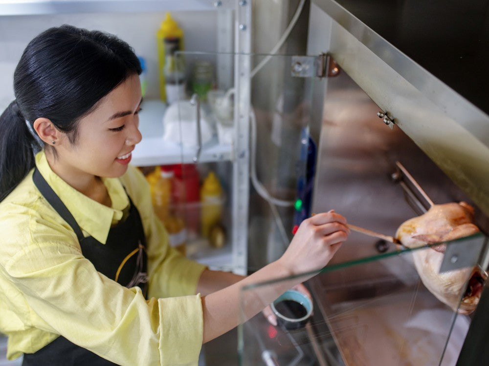A kitchen worker basting a whole chicken on a countertop rotisserie in a food service kitchen