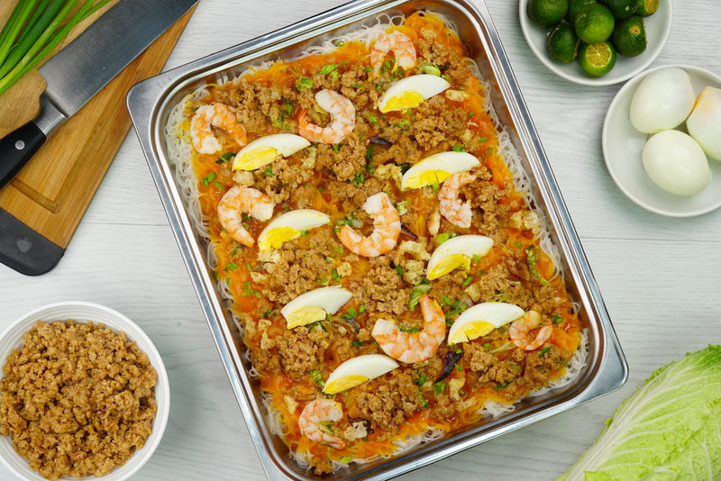 Pancit palabok in a food service tray topped with shrimp, egg wedges, and crushed chicharon