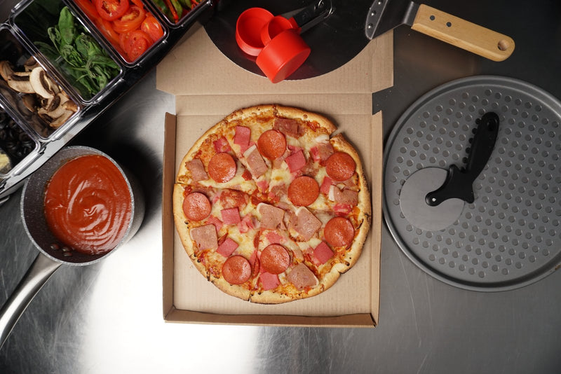 All-meat pizza in an open box with pepperoni and ham, styled with a food service mise en place