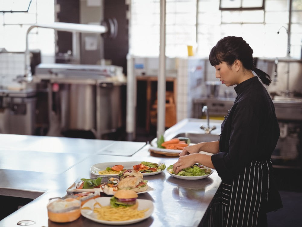 A cook plating salads and sandwiches on a stainless prep counter in a commercial kitchen